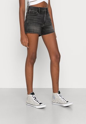 Person trägt schwarze Jeansshorts, weißes Crop-Top und weiße High-Top-Sneaker mit schwarzen Flecken, steht vor einem schlichten hellen Hintergrund.