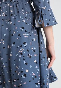 Sininen mekko, jossa on abstrakti vaaleanpunainen, vaaleansininen ja musta kuvio. Kellohiha, jossa on nauhat. Kevyt kangas, rento malli, polvipituinen helma.