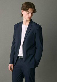 Blazer navy con rever a taglio e due bottoni, abbinato a una maglietta bianca semplice. Tessuto liscio, design aderente e tasca laterale singola.