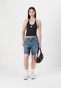 Musta halterneck crop top valkoisilla yksityiskohdilla ja logolla, yhdistettynä sinisiin farkkushortsit bermuda-mallissa. Malli pitää käsissään tummaa olkalaukkua ja käyttää valkoisia tennareita.