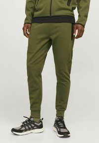 Olive green jogger pants made of soft material with tapered leg design, zippered side pockets, and contrasting black waistband.