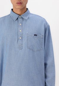 Cămașă din denim bleu deschis, cu placket cu nasturi, mâneci lungi, buzunar tip patch și logo brodat "GAP" pe piept.