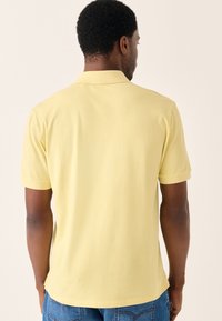 Gul poloshirt med blød tekstur, korte ærmer og klassisk krave. Båret med blå denimjeans, der viser en afslappet pasform.