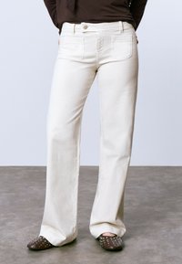 Jeans de pierna ancha color crema con bolsillos frontales y traseros, que cuentan con un cierre de botón y una textura suave. Combinando con mule marrones con tachuelas.