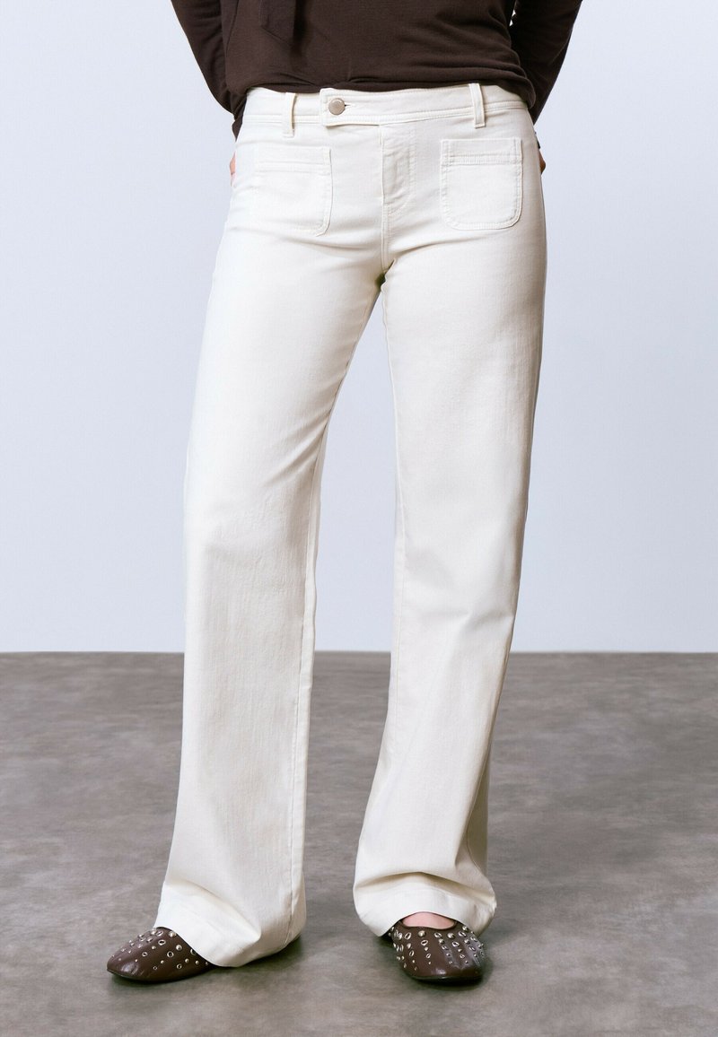 Jeans de pierna ancha color crema con bolsillos frontales y traseros, que cuentan con un cierre de botón y una textura suave. Combinando con mule marrones con tachuelas.