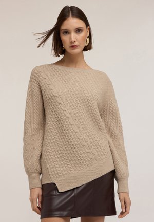 Beżowy sweter dzianinowy z teksturowymi wzorami, asymetrycznym dołem i ściągaczami, połączony z ciemnobrązową skórzaną spódnicą.