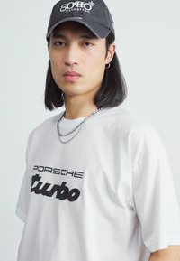 Balta T-kreklu ar īsām piedurknēm, uz kura ir melns uzraksts "PORSCHE turbo". To papildina tumša cepure ar logo "SOHO MANHATTAN" un slāņoti sudraba ķēdītes.