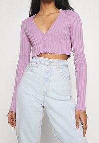 Kvinna i en kortad, ribbad, lavendelfärgad knappad cardigan och högmidjade, ljusblå jeans med avslappnad passform.