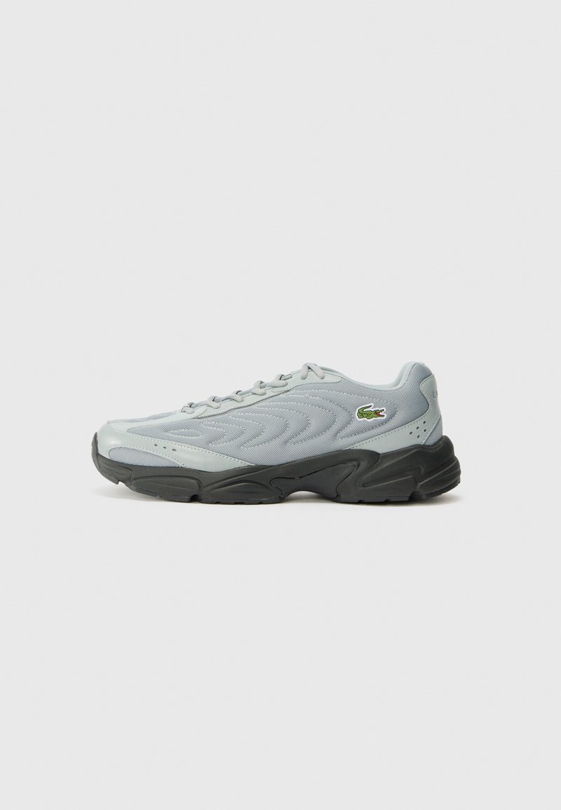 Grijze sportieve schoen met een gestructureerde bovenkant, zwarte rubberen zool en vetersluiting. Bevat een klein groen logo aan de zijkant.