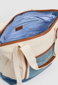 Plátěná taška s hnědými koženými akcenty. Světle béžový exteriér, spodní část z modrého denimu. Interiér má modro-bílé pruhované podšívku s kapsou na zip.