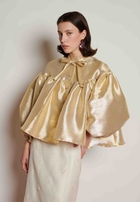 Μεταλλική χρυσή κάπα με φουσκωτά μανίκια, διαθέτει δέσιμο με φιόγκο και διακοσμητικά μαργαριτάρια. Συνδυάζεται με κρεμ φούστα διακοσμημένη με κεντήματα λουλουδιών.