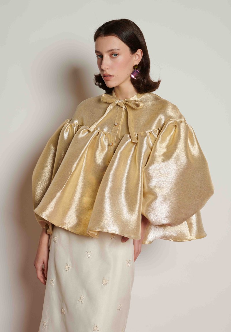 Μεταλλική χρυσή κάπα με φουσκωτά μανίκια, διαθέτει δέσιμο με φιόγκο και διακοσμητικά μαργαριτάρια. Συνδυάζεται με κρεμ φούστα διακοσμημένη με κεντήματα λουλουδιών.