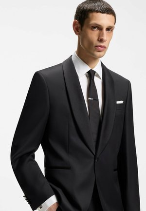 Man draagt een zwart smoking met satijnen revers, wit overhemd, zwarte stropdas met dasspeld en een wit pochet tegen een effen achtergrond.