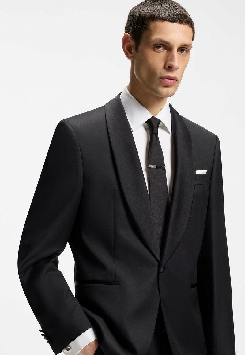 Mężczyzna ubrany w czarny smoking z satynowymi klapami, białą koszulę, czarny krawat z klipsem oraz białą poszetkę na jednolitym tle.