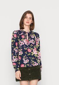 Blusa floral azul-marinho com mangas longas, apresentando flores cor-de-rosa e verdes, uma abertura em forma de buraco de fechadura com laços, e um ajuste solto. Combinada com uma saia de veludo cotelê verde.