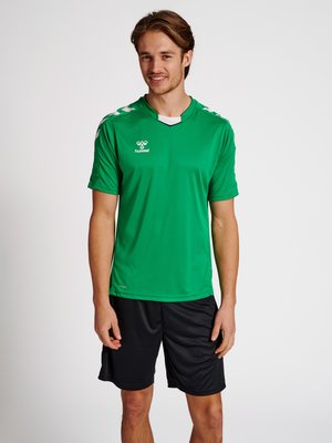 Ung mand smiler, iført grøn kortærmet sportstrøje og sorte træningsshorts, står foran en ensfarvet lys baggrund.