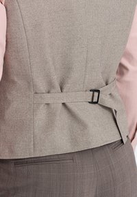 Chaleco gris ajustado con tejido texturizado, que presenta un detalle de hebilla negra en la cintura, combinado con una camisa rosa clara y pantalones marrones estampados.