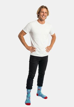 Lächelnder Mann in weißem T-Shirt und schwarzer Jeans steht mit den Händen in der Hüfte, trägt leuchtend blaue Socken mit roten Seestern- und Oktopusmustern.