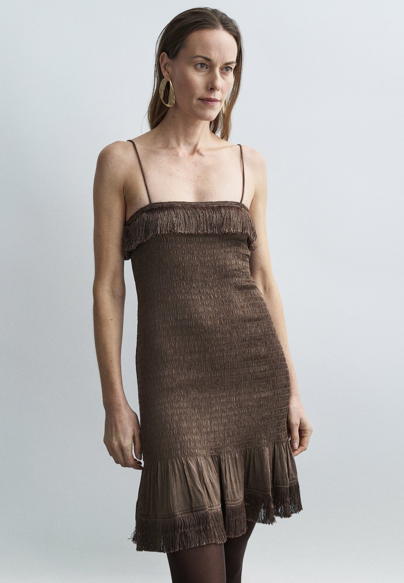 Brun smokket minikjole med spaghetti-stropper, med frynsete øvre kant og en volangkant nederst. Stoffet har et teksturert, vevd utseende.