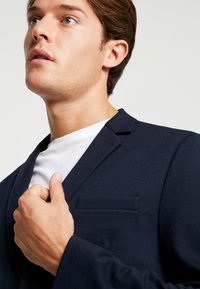 Blazer azul marino con solapa estructurada de muesca y un bolsillo en el pecho, usado sobre una camiseta blanca. Tejido suave con un ajuste entallado.