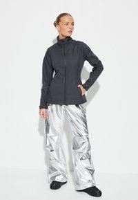 Chaqueta softshell negra, ajustada, con cuello alto y cierre de cremallera. Combinada con pantalones cargo plateados brillantes de pierna ancha y calzado negro.