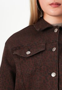 Kvinnen har på seg en brun og svart leopardmønstret jakke med knapper i metall og en beige topp under.