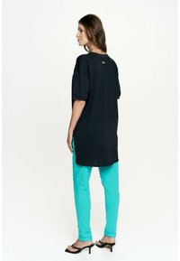Czarna, oversize koszulka z krótkim rękawem, w zestawieniu z turkusowymi, zwężającymi się spodniami i czarnymi sandałami na obcasie, widok z tyłu.