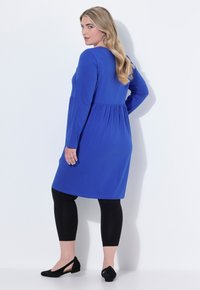 Modrá dlouhá šatová šaty s nařaseným pasem, spárované s černými legínami a černými plochými botami. Hladká tkanina a jednoduchý design.