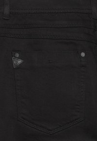 Close-up af baglommen på sorte denimjeans med forstærket syning og to metalknappenåle i hjørnerne øverst.