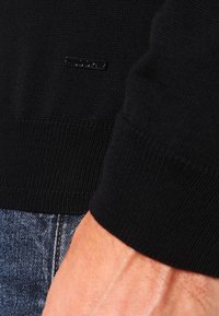 Maglione nero lavorato a maglia con orlo e polsini a coste, dotato di una piccola etichetta logo lucida sul lato anteriore vicino all'orlo.