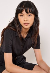 Robe chemise noire à manches courtes avec fermeture à boutons, poche poitrine unique et ceinture en tissu. Texture lisse, coupe décontractée et longueur genoux.
