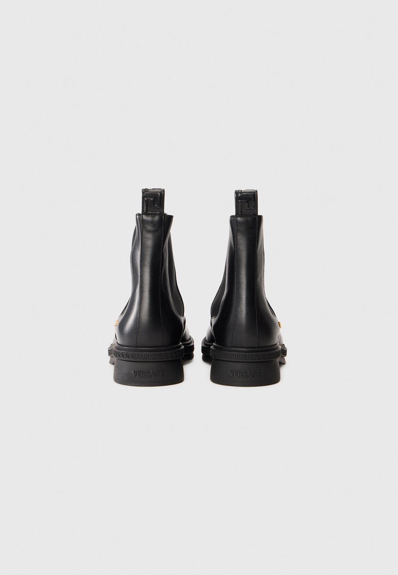 Stivali alla caviglia in pelle nera con linguette di estrazione, motivo a chiave greca in rilievo sui talloni e marchio "Versace" sui bordi delle suole.