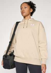 Beige hoodie med känguruficka, med ribbad mudd och nederkant, samt en vit logotyp på bröstet. Svart axelväska med nät detaljer.