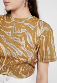Camiseta de manga corta y cuello redondo en color verde oliva, con un patrón abstracto ondulado en tonos de azul, durazno y blanco. Textura de tela suave.