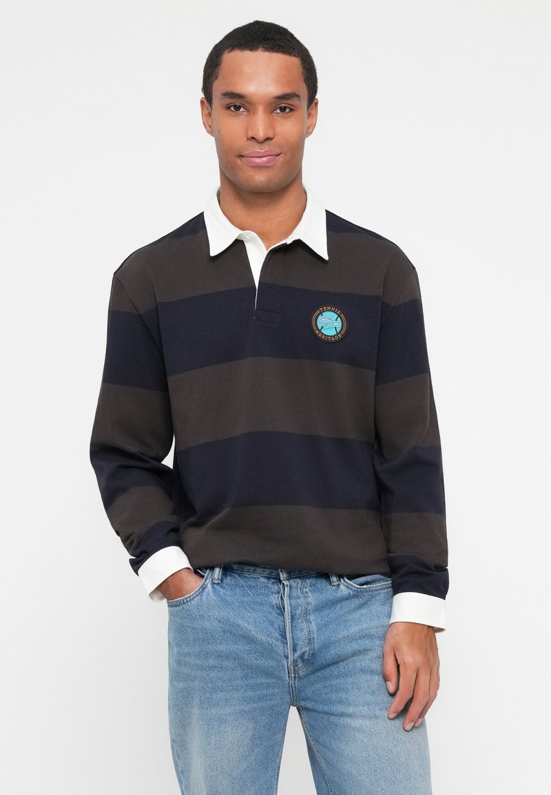 Langærmet polo shirt med marineblå og mørkegrå vandrette striber, hvid krave og broderet logo på venstre bryst. Bæres med lyseblå jeans.