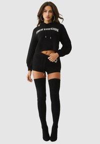 Schwarzer, kurz geschnittener Strick-Hoodie mit dem Schriftzug "TRUE RELIGION", kombiniert mit schwarzen Strickshorts und schwarzen Overknee-Stiefeln aus Wildleder. Durchgehend glatte Textur.