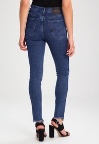 Jeans de denim oscuro y ajustado con una cintura alta, que presentan bolsillos traseros y dobladillos deshilachados, combinados con sandalias de tacón alto negras.