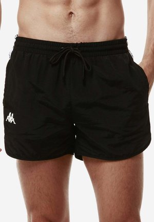 Torace maschile che indossa pantaloncini neri Kappa con coulisse in vita e logo sulla gamba sinistra, in piedi con le mani nelle tasche su sfondo bianco.