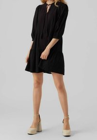 Robe noire courte avec une coupe décontractée, manches longues bouffantes, corsage plissé et jupe à volants. Présente un lien au niveau du décolleté avec des franges et une texture douce.