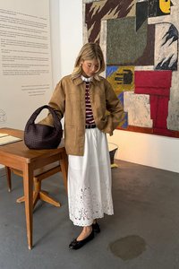 Brązowa zamszowa kurtka z dużymi kieszeniami, bordowa koszula w paski, biała spódnica z koronkowym dołem, czarne płaskie buty oraz torebka z fakturowanej plecionki. Tło z motywem artystycznym.