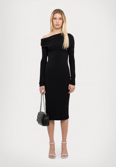 Rochie neagră, cu umerii la vedere, dintr-un material lucios, cu un design ajustat, de lungime medie până la glezne, asortată cu o geantă mică neagră și tocuri albe.