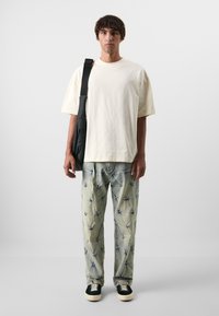 Crème oversized t-shirt, lichtgepatroonde denim broek met blauwe accenten, zwarte sneakers. Model houdt een zwarte schoudertas vast. Minimalistische achtergrond.