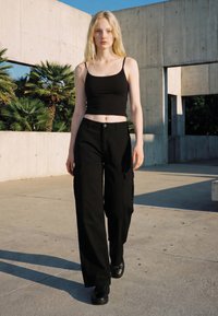 Lyshåret kvinde, der går udendørs iført en sort crop top, vidde cargobukser og sorte sneakers mod beton og palmer.