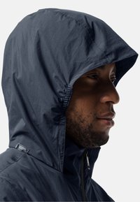 Veste à capuche marine avec une capuche élastique froncée, en tissu texturé et à fermeture éclair. Elle dispose d'un détail d'étiquette sur le côté, près de l'épaule.