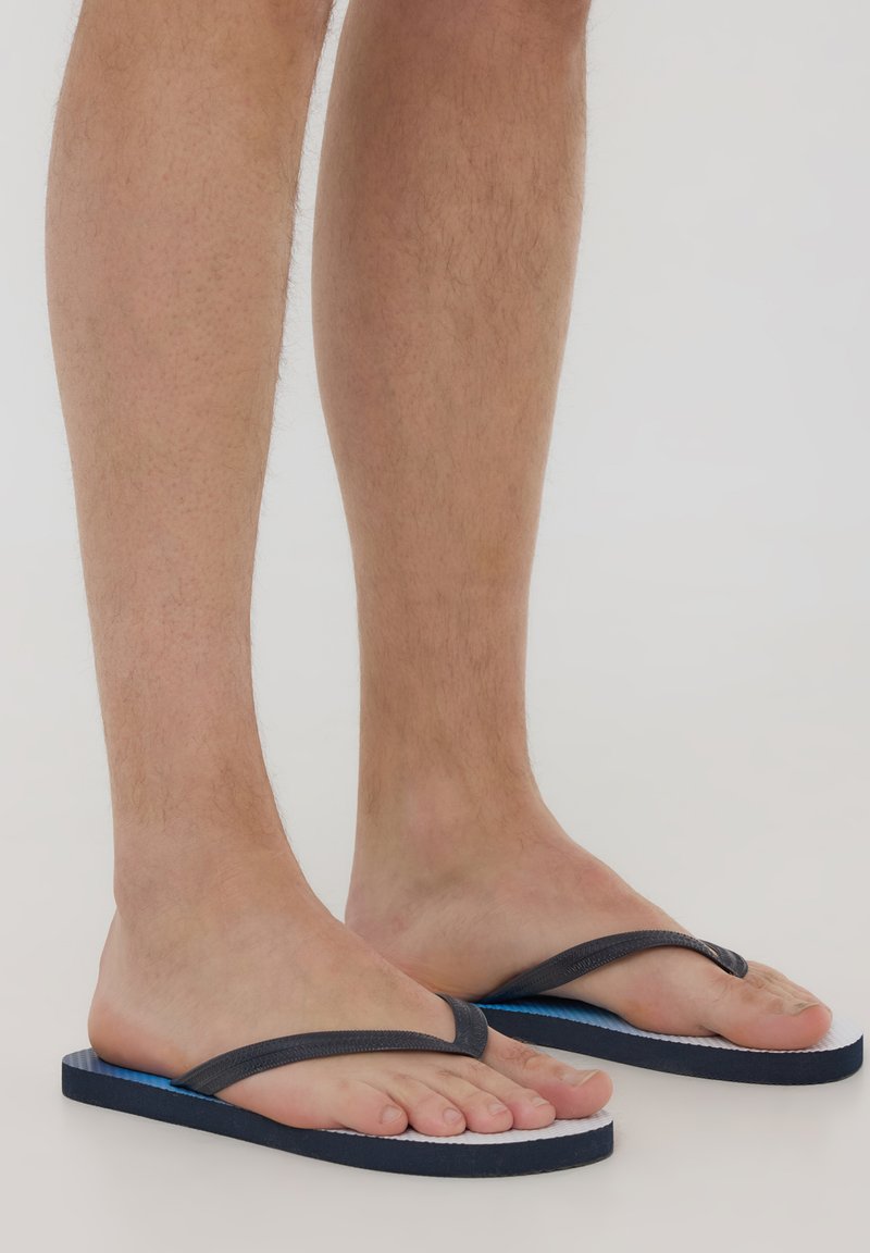 Infradito grigie scure con una fascia testurizzata, suola blu e bianca, design piatto e materiale in gomma. Indossate a piedi nudi.