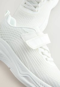 Chaussures de sport blanches avec une tige en mesh texturé, une sangle ajustable et un système de laçage. Conçues avec une semelle rembourrée pour le confort.