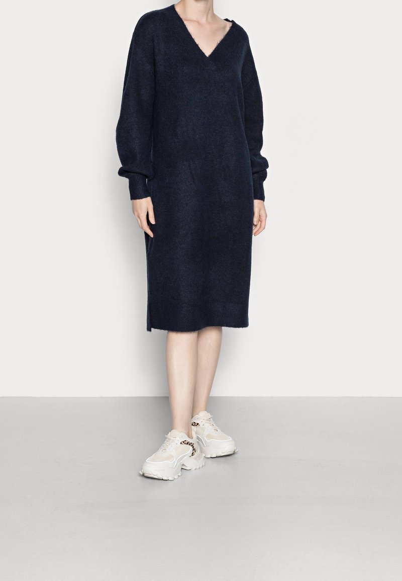 Personne debout portant une longue robe-pull bleu marine foncé à col en V et des baskets blanches épaisses avec des détails imprimés léopard.