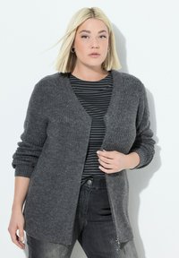 Blondýnka oblečená v tmavě šedém pleteném kardiganu přes černý a šedý pruhovaný top, doplněná o tmavé džíny s lehkým opráním.