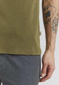 Olijfgroene katoenen t-shirt met een gestructureerd oppervlak en een label aan de zoom. Grijze broek gedeeltelijk zichtbaar aan de onderkant van de afbeelding.