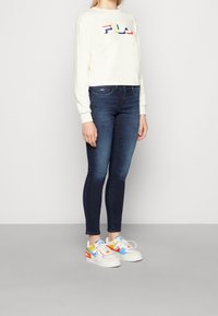 Persona che indossa una felpa bianca FILA, jeans skinny blu scuro e sneakers bianche con accenti blu, gialli e rossi, in piedi su un pavimento grigio.